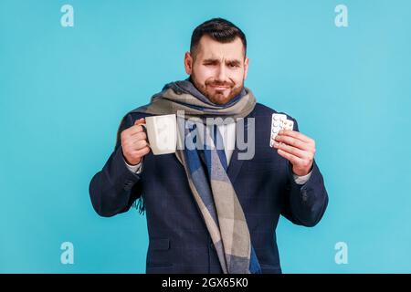 Portrait of sad sick business wearing official style suit and wrapped in scarf, treating with pills and warm tea, being upset, feeling unwell. Indoor studio shot isolated on blue background. Stock Photo