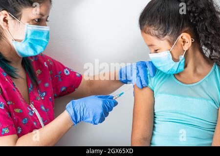 children's nurse injecting arm of little brown girl, while looking at her face. doctor's hands with rubber gloves injecting covid-19 or flu vaccine. m Stock Photo