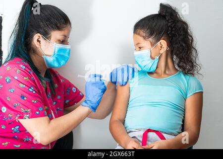 children's nurse injecting arm of little brown girl. doctor's hands with rubber gloves injecting covid-19 or flu vaccine. girl sitting while being vac Stock Photo