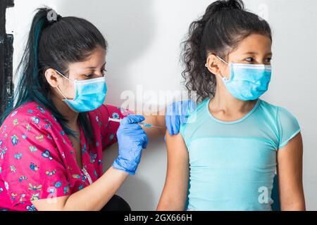 children's nurse injecting arm of little brown girl. girl looking away, doctor's hands with rubber gloves injecting covid-19 or flu vaccine. medical c Stock Photo