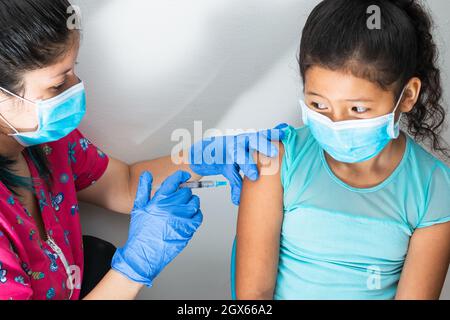 children's nurse injecting arm of little brown girl. frightened girl with scary face, doctor's hands with rubber gloves injecting covid-19 vaccine. fl Stock Photo