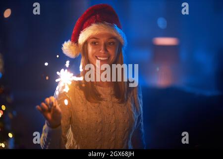 Happy caucasian woman in santa hat standing at dark living room with burning sparklers in hands. Pretty blonde celebrating new year of christmas at home. Concept of winter holidays. Stock Photo