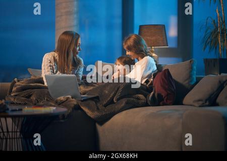 Happy caucasian mother and her two cute daughters using modern laptop for watching funny videos during evening time at home. Family time and technology concept. Stock Photo