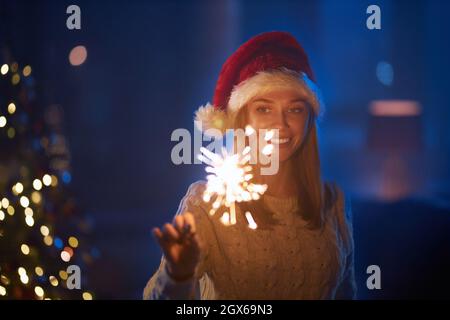 Cheerful young woman in red santa hat standing at dark living room and holding sparklers. Charming caucasian blonde enjoying winter holidays at home. Celebration concept.  Stock Photo