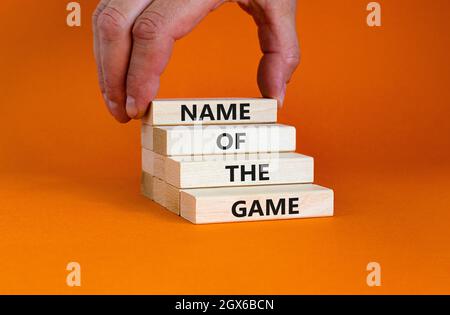 Name of the game symbol. Wooden blocks with concept words 'Name of the game'. Businessman hand. Beautiful orange background. Business and name of the Stock Photo