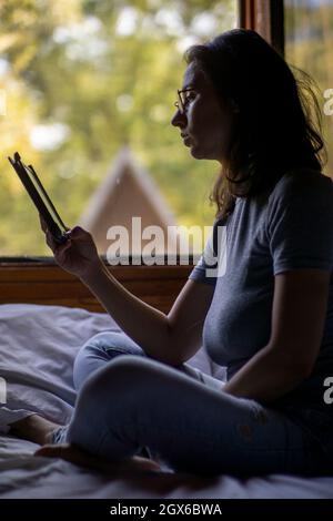 Attractive girl sitting near window of wooden bungalow with autumn trees view, reading on a ebook. Young woman relaxing in luxury villa on family summ Stock Photo