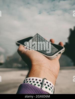 PARIS, FRANCE - Aug 28, 2021: A vertical shot of a hand holding iPhone with the Louvre Museum coming out from inside. Photoshop edited Stock Photo