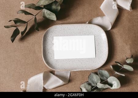 Boho wedding, birthday stationery. Blank business card, invitation mockup with dry eucalyptus branches on dotted textured ceramic plate. Beige grunge Stock Photo