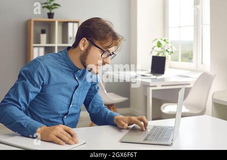 Young man sitting at desk, using laptop, studying new information online and taking notes Stock Photo