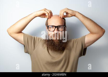 Male touching his head. problem with hair, suffering alopecia, needs trichologist consultation, bald bearded man wearing T-shirt and glasses. Indoor studio shot isolated on gray background. Stock Photo