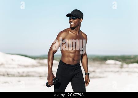 Portrait of confident African American male athlete, muscular athlete standing on mountain road at sunset, with bottle of drinking water, outdoor spor Stock Photo