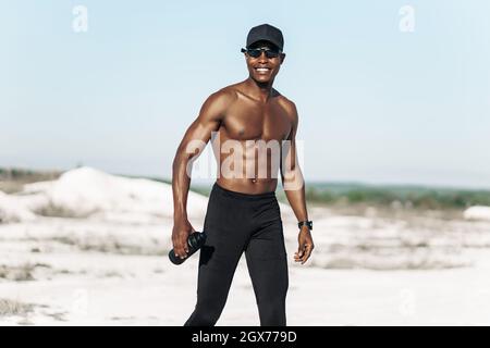 Portrait of confident African American male athlete, muscular athlete standing on mountain road at sunset, with bottle of drinking water, outdoor spor Stock Photo