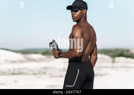 Portrait of confident African American male athlete, muscular athlete standing on mountain road at sunset, with bottle of drinking water, outdoor spor Stock Photo