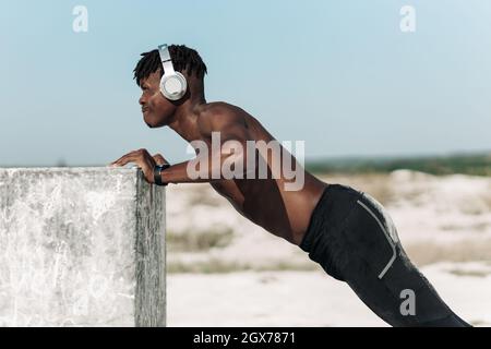 Sporty african black man, wearing wireless headphones, doing push-ups outdoors, man doing push-up exercises leaning on beaton cube Stock Photo