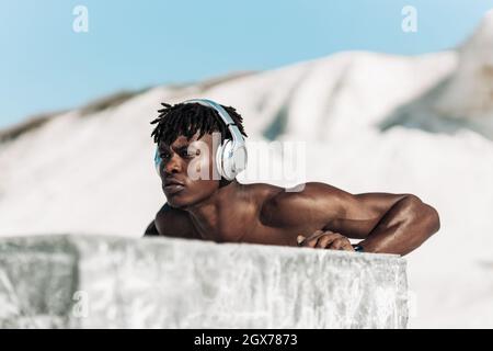 Sporty african black man, wearing wireless headphones, doing push-ups outdoors, man doing push-up exercises leaning on beaton cube Stock Photo