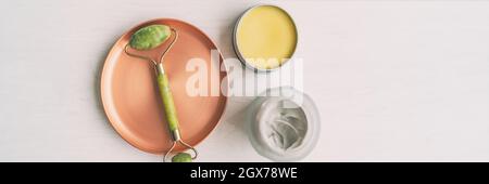 Facial beauty product jade face roller top view of massage tool with jars of shea butter and natural clay banner panorama. Skincare wellness cosmetic Stock Photo