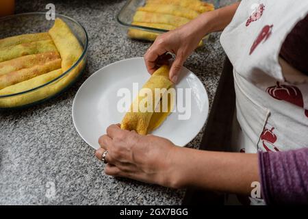 Woman in an apron rolling the stuffed pancake for preparing delicious homemade Cannelloni Stock Photo