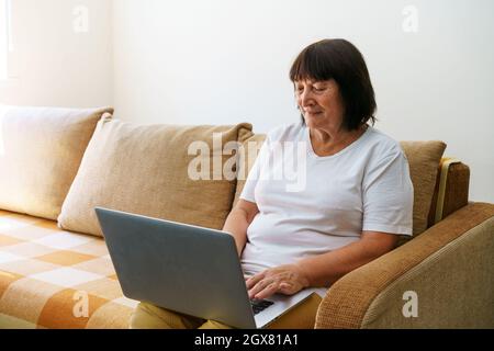 Elderly woman smiling 60s, sitting at home on couch, holding laptop. Senior happy business woman using computer technology, work, conversation, pastime on social networks internet, e-commerce. Stock Photo