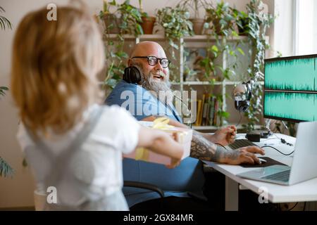 Cute little girl gives present to surprised dad working on computer in light room Stock Photo