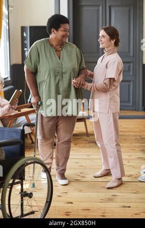 Vertical full length portrait of smiling young woman helping seniors in modern retirement home Stock Photo