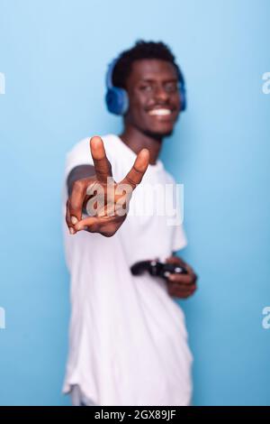 African american man showing peace sign at camera while wearing headphones and holding controller. Black person feeling joyful and doing gesture with hand while standing over background Stock Photo