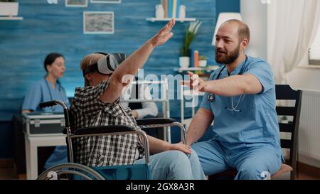 Retired woman using vr glasses and nurse assisting, explaining virtual reality in nursing facility. Senior patient sitting in wheelchair wearing headset at checkup with medical specialist Stock Photo