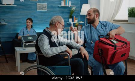 Medical assistant using thermometer for symptoms checkup with disabled man in nursing home. Nurse measuring temperature of senior patient in wheelchair for healthcare examination. Stock Photo