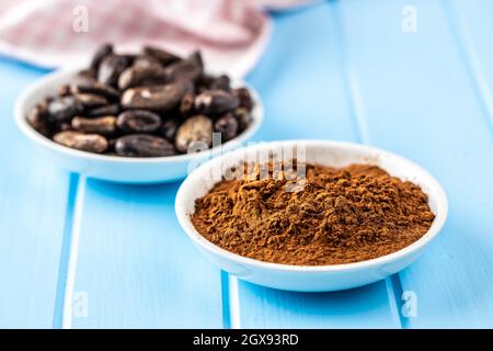 Roasted cocoa beans and cocoa powder in bowl on blue table. Stock Photo