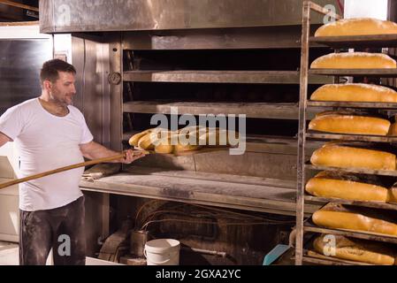 bakery worker taking out freshly baked breads with shovel from the professional oven at the manufacturing Stock Photo