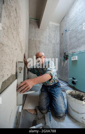 experienced senior construction worker using level tool while installing big ceramic tiles on interior concrete walls Stock Photo
