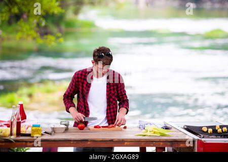 young man cutting vegetables for salad or barbecue grill during outdoor french dinner party near the river on beautiful summer evening in nature Stock Photo