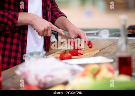 young man cutting vegetables for salad or barbecue grill during outdoor french dinner party near the river on beautiful summer evening in nature Stock Photo