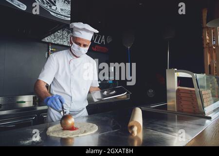 Skilled chef preparing traditional italian pizza  in interior of modern restaurant kitchen with special wood-fired oven. Wearing protective medical fa Stock Photo