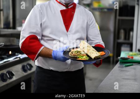chef cook wearing face protective medical mask for protection from corona virus disease with plate of authentic sandwich at professional restaurant ki Stock Photo