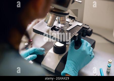 Lab assistant in latex gloves looks into microscope at sample in laboratory closeup Stock Photo