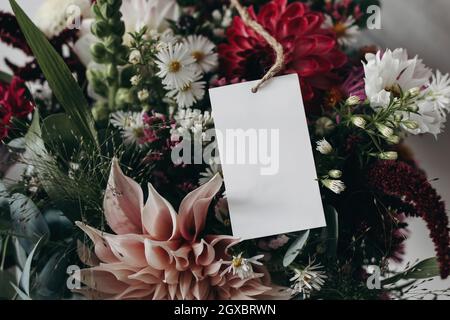 Closeup of blank paper card, gift tag label on colorful floral. Moody autumn wedding, birthday mockup scene. Pink and burgundy dahlia , cosmos and Stock Photo