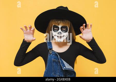 Portrait girl child with Halloween makeup mask, wears black hat says Boo laughs, hands raised, spooky looking at camera, posing on yellow wall Stock Photo