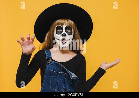 Happy Halloween concept. Shocked little girl child with spooky makeup wears big black hat showing empty copy space on open hand palm Stock Photo