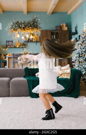 A cute girl holds a soft toy in her hands and dances with it, spinning in a room decorated for Christmas, against the background of a Christmas tree Stock Photo