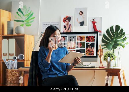 Skilled young Asian designer sitting against computer and holding tablet while discussing design of web project by phone Stock Photo