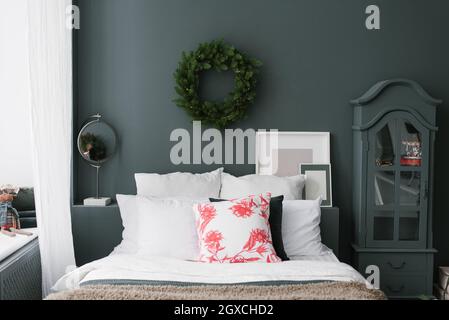 Pillows on the bed, above which a Christmas wreath hangs on a dark wall. Chest of drawers, mirror and mockups photo frames Stock Photo