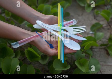 Hands holding used plastic dishes and straws on the beach. Beautiful green grass on the background. Environmental Problem. Single-use plastic waste  Stock Photo