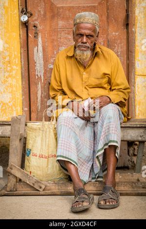 INDIA, BANGLADESH - DECEMBER 6, 2015: Senior ethnic elderly male in traditional clothes sitting on shabby weathered wooden door holding money and look Stock Photo