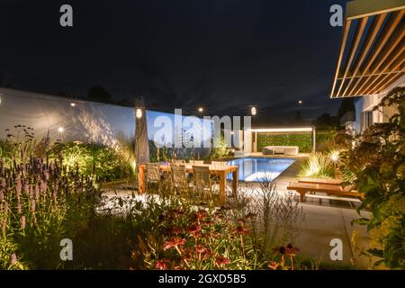Spacious backyard of contemporary villa with wooden table and sun loungers and green plants placed near swimming pool against cloudless night sky Stock Photo