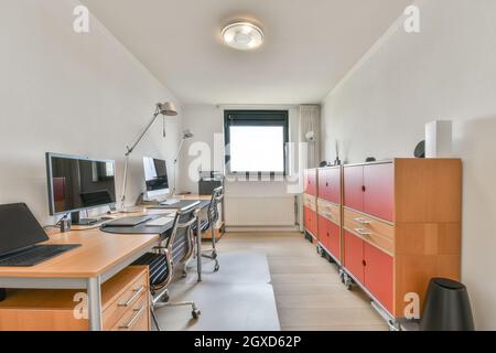 Desktop computers and laptop against armchairs and cabinets on parquet under lamp in contemporary workspace Stock Photo