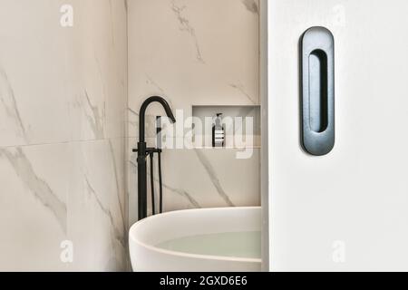 Opened door to light bathroom with marble walls and black bathroom accessories above soaking tub filled with water Stock Photo