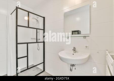Minimalist design of white bathroom with sink under mirror hanging on tiled wall near glass shower cabin in apartment Stock Photo