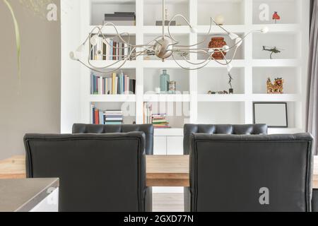 Empty wooden table and black leather chairs placed in stylish room against white shelves with books and decorative elements Stock Photo