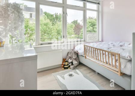 Toys placed on floor with carpet near comfortable bed in children bedroom with large window in daytime Stock Photo