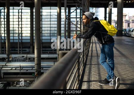 Side view of ethnic male traveler with backpack standing near railing on passage above trains at railway station Stock Photo